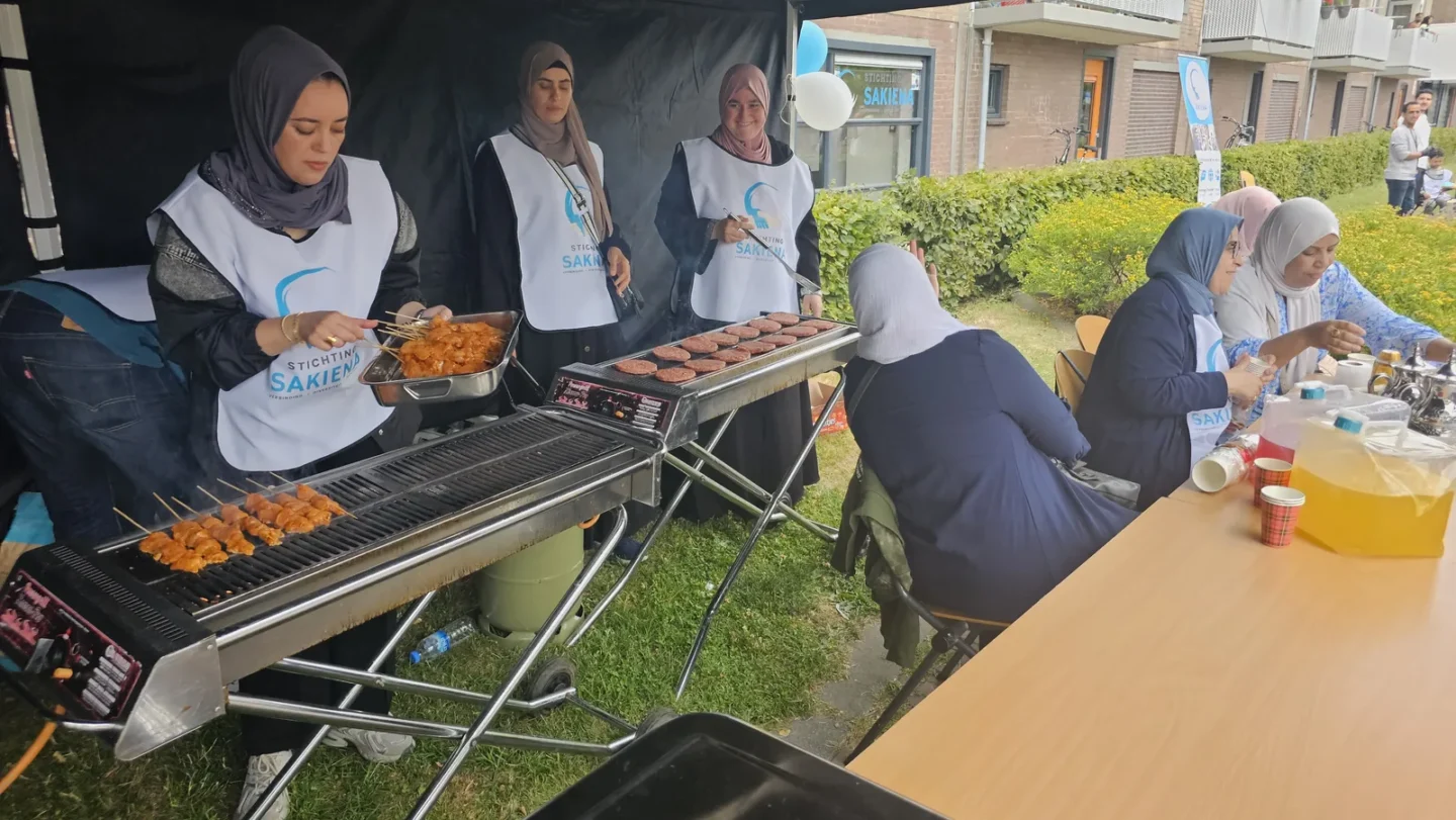 Drie vrijwilligers in Sakiena-hesje grillen kipspiesjes tijdens het buurtfeest