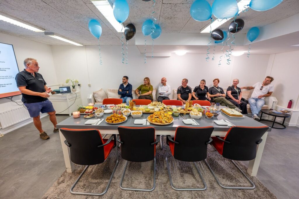 Man geeft een presentatie aan genodigden die rond een gedekte tafel zitten, met ballonnen aan het plafond