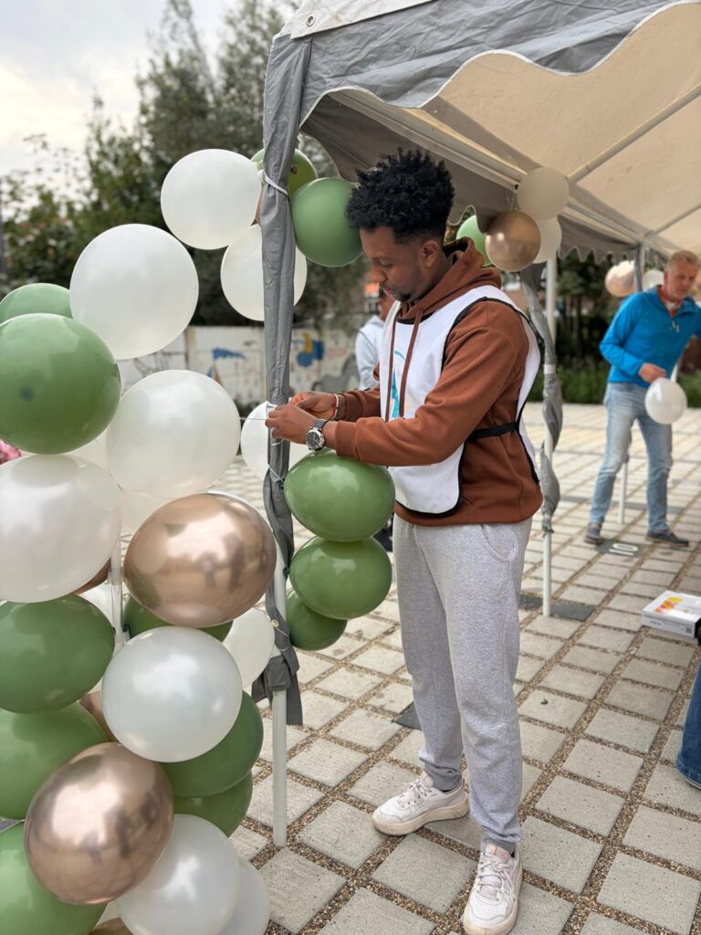Jongeman in een hesje van Stichting Sakiena bindt groene, witte en gouden ballonnen aan een tentpaal