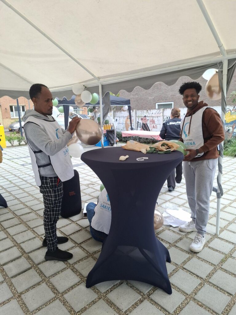 Twee mannen in een hesje van Stichting Sakiena maken ballonnen klaar aan een statafel onder een partytent