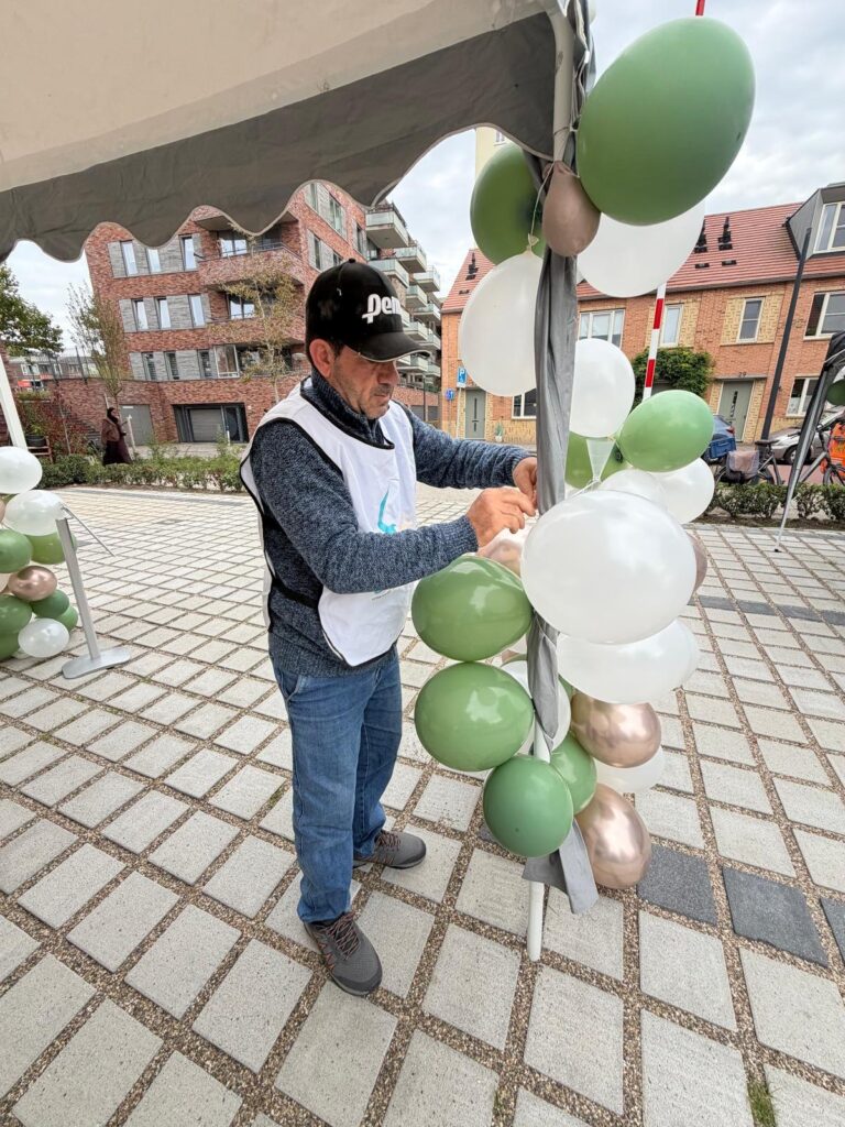 Man in een hesje van Stichting Sakiena maakt een pilaar van ballonnen vast aan een partytent, met nieuwbouw op de achtergrond