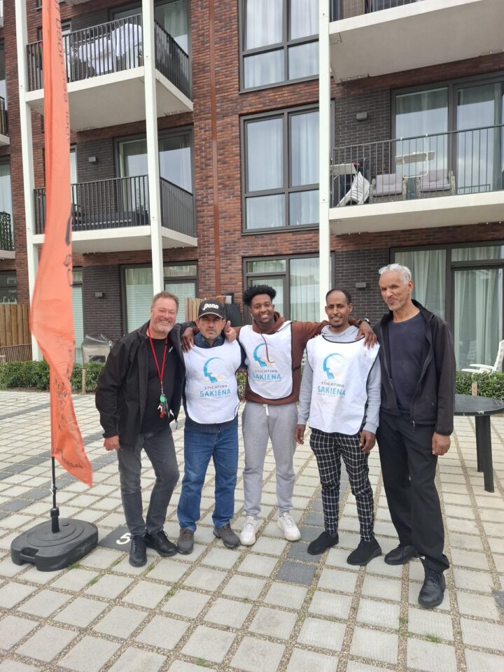 Vijf mannen staan samen op het plein voor de nieuwe huurappartementen van De Sleutels. Drie van hen dragen een hesje van Stichting Sakiena.