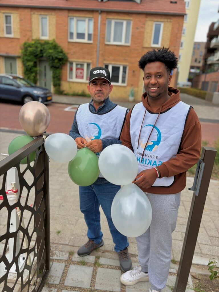 Twee mannen in een hesje van Stichting Sakiena met ballonnen bij een tuinhek