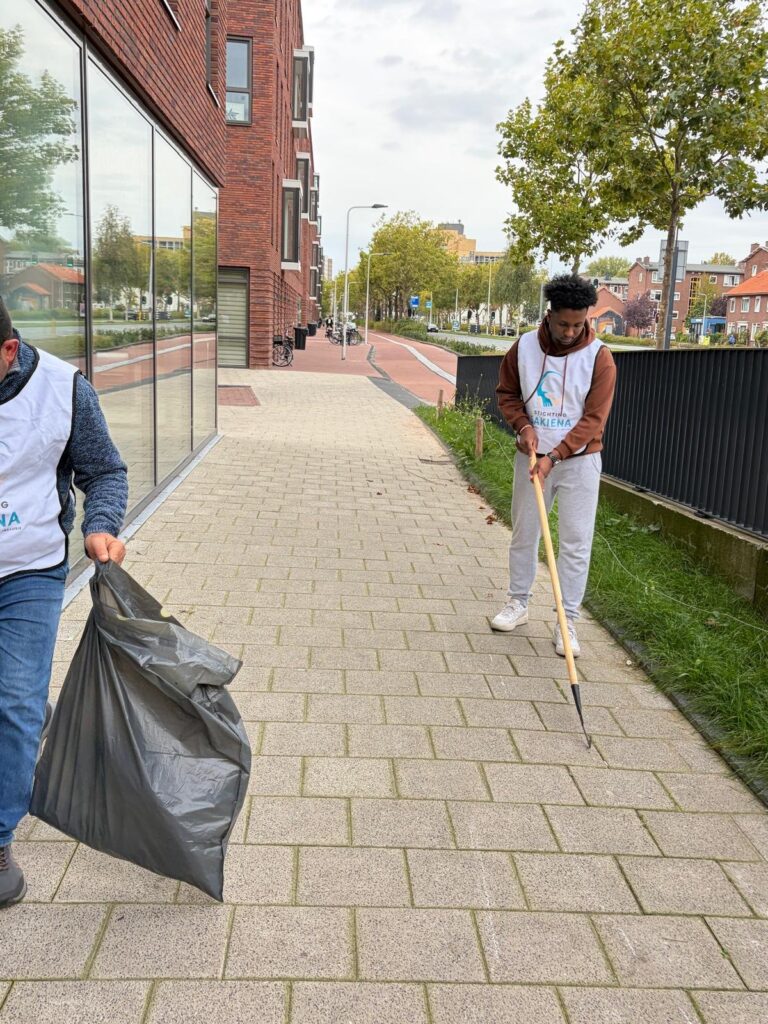Twee mannen in een hesje van Stichting Sakiena ruimen zwerfvuil op langs een fietspad, met een grijper en een vuilniszak