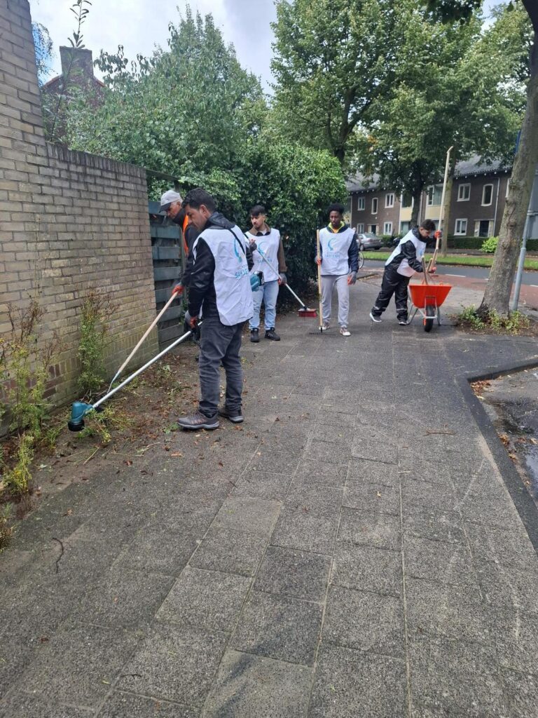 Deelnemers in hesjes van Stichting Sakiena verwijderen onkruid langs een tuinmuur, met een kruiwagen op de stoep