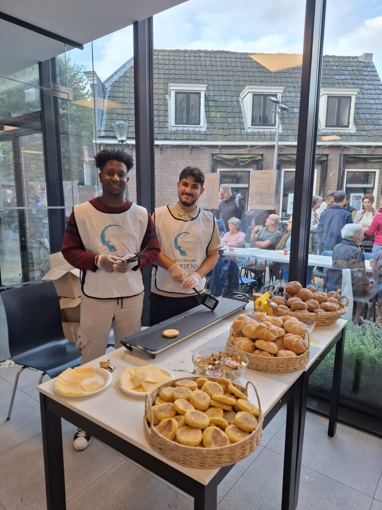 Twee vrijwilligers in hesjes van Stichting Sakiena bij de broodtafel, met uitzicht op de binnenplaats