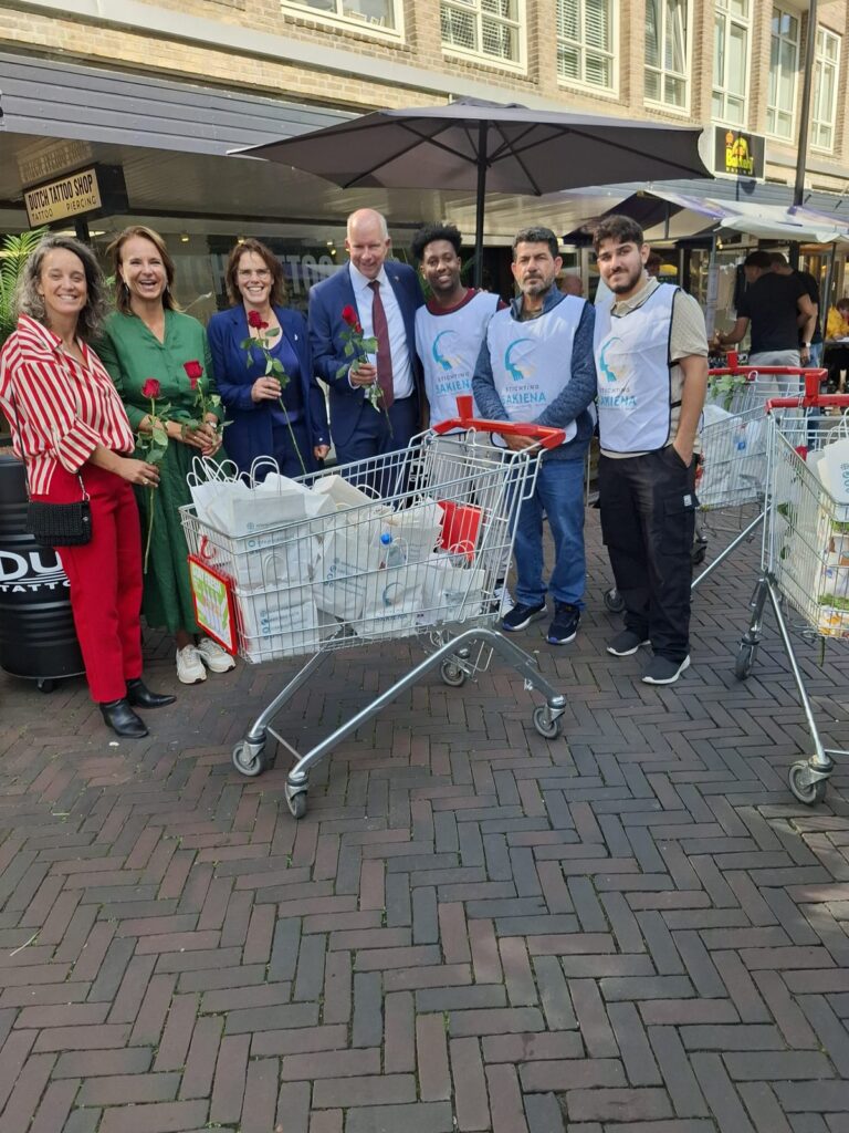 Groepsfoto bij twee winkelwagens vol Sakiena-tassen, met rode rozen in de hand
