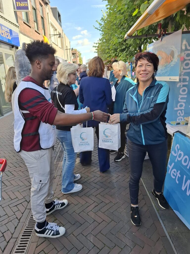 Vrijwilliger in een hesje van Stichting Sakiena geeft een tasje met goodies aan een vrouw op straat