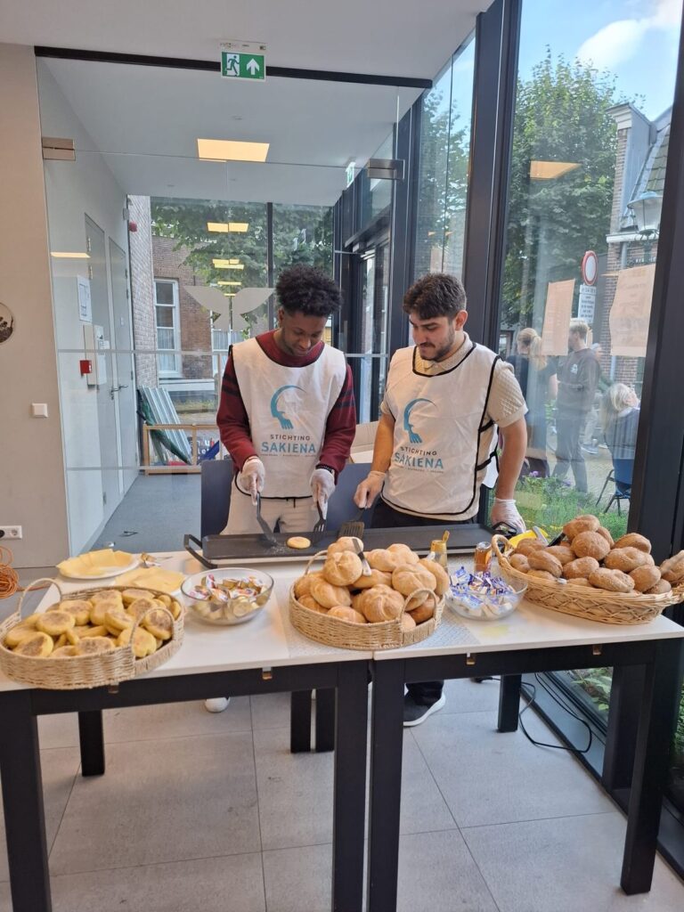 Twee jonge vrijwilligers in hesjes van Stichting Sakiena achter de bakplaat aan de ontbijttafel