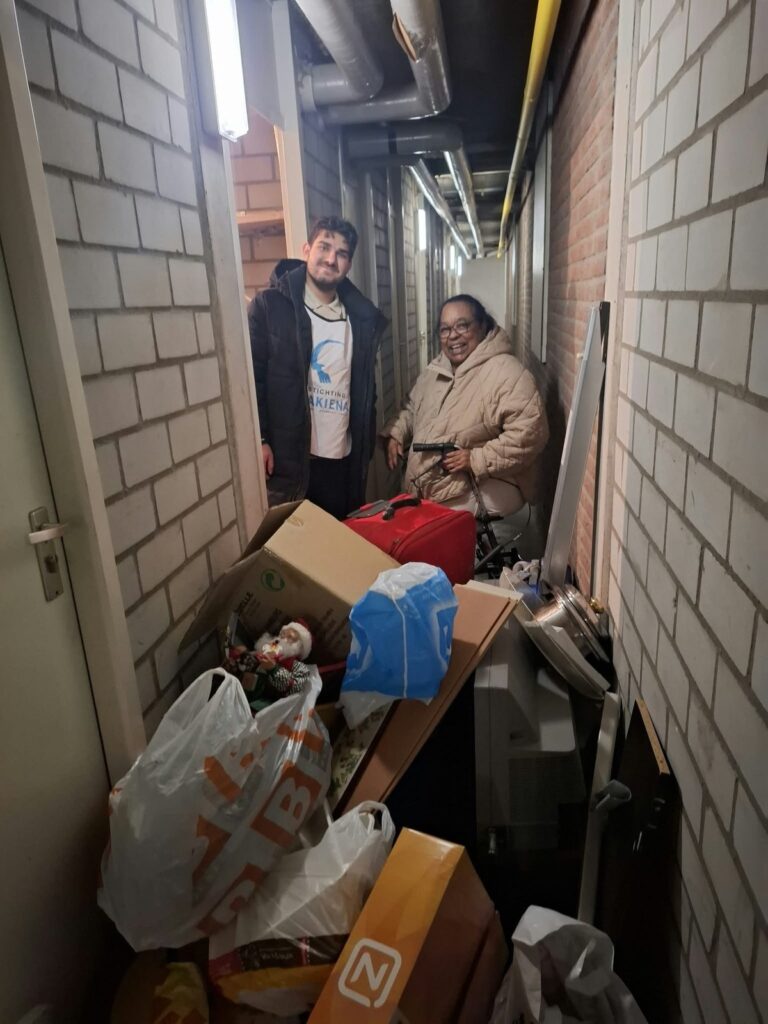 Een lachende oudere bewoonster met een rollator staat in een smalle gang bij de bergingen, naast een deelnemer in een wit hesje van Stichting Sakiena. Voor hen staan dozen, tassen en een rode koffer.