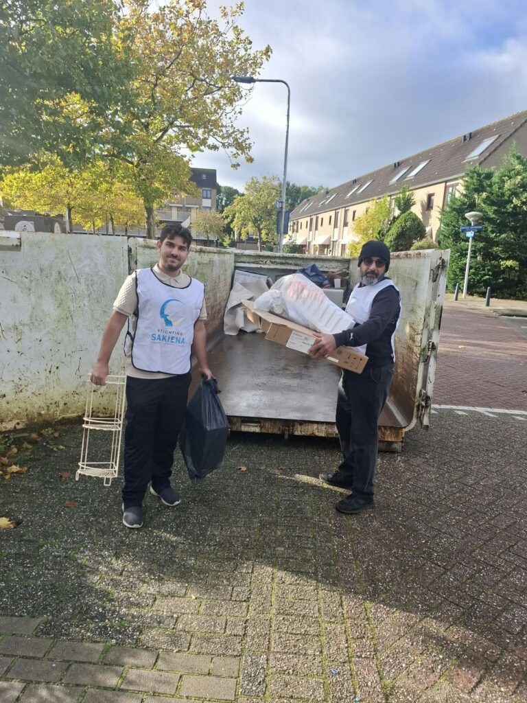 Twee deelnemers in witte hesjes van Stichting Sakiena staan bij een grote, gevulde grofvuilcontainer in een woonstraat. Een van hen draagt een doos, de ander een vuilniszak.