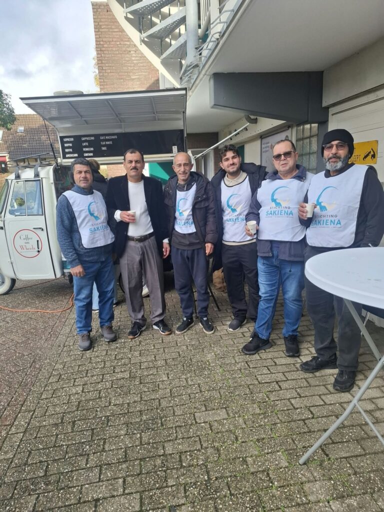 Zes deelnemers, de meesten in witte hesjes van Stichting Sakiena, staan samen voor de koffiekar met een kopje koffie in de hand.