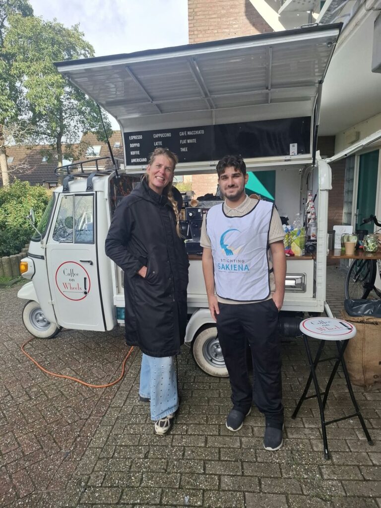 Een vrouw en een deelnemer in een wit hesje van Stichting Sakiena staan naast een kleine koffiekar op straat. Op de kar staat een menubord met koffiesoorten.
