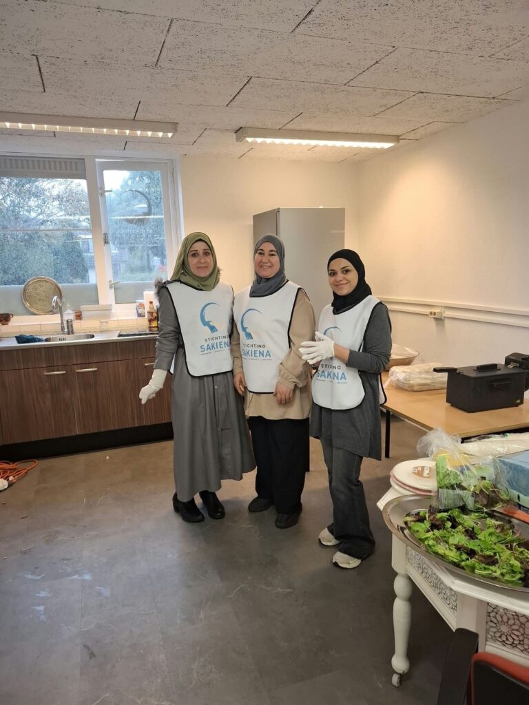 Drie vrouwen van de kookgroep in witte schorten van Stichting Sakiena staan glimlachend in een keuken. Op tafel staat een schaal salade.
