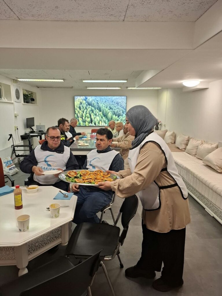 Een vrouw van de kookgroep brengt een grote schaal eten naar een tafel. Aan de tafel zitten bewoners en deelnemers in witte hesjes, achter hen een groot scherm met een bos.