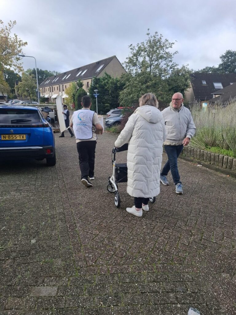 Een deelnemer in een wit hesje loopt over straat naar de container. Een oudere bewoonster met een rollator en een bewoner lopen met hem mee.