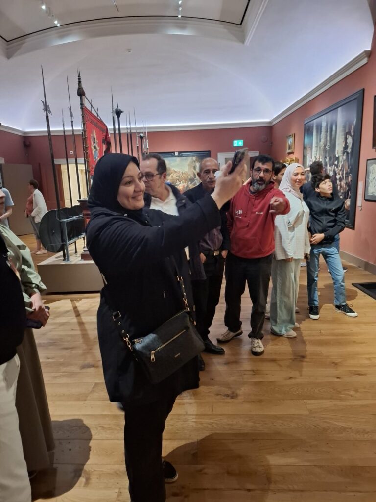 Een deelneemster maakt lachend een foto met haar telefoon in het museum. Achter haar staat de groep, met oude lansen en een rood vaandel op de achtergrond.