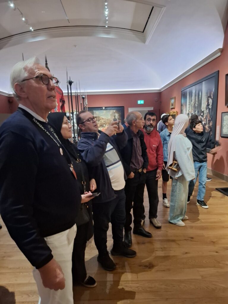 Deelnemers van jong tot oud kijken omhoog naar de schilderijen aan de rode muren van het museum. Een van hen maakt een foto met zijn telefoon.