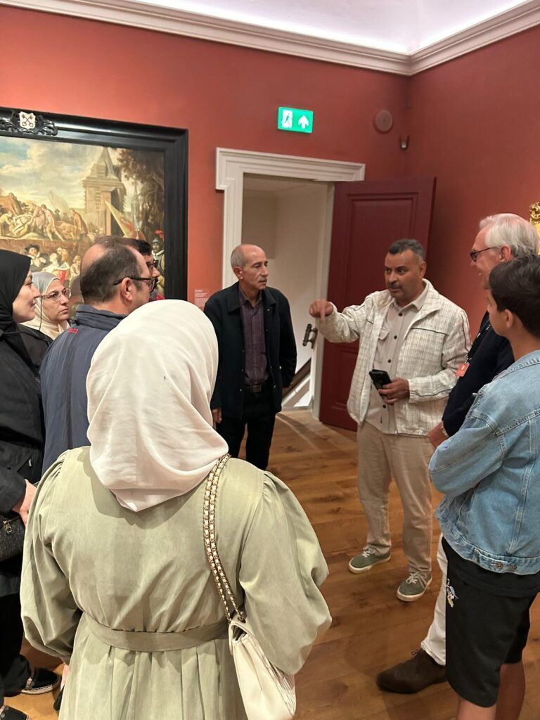 De groep staat in een kring in een zaal met rode muren en luistert naar uitleg. Links hangt een groot oud schilderij.
