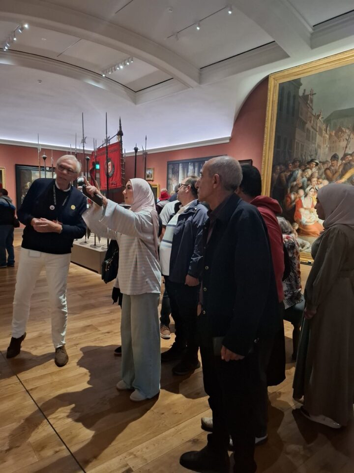 Een groep deelnemers van Stichting Sakiena krijgt uitleg in een zaal van Museum De Lakenhal. Een vrouw wijst naar boven, naast oude lansen, een rood vaandel en een groot schilderij in een gouden lijst.