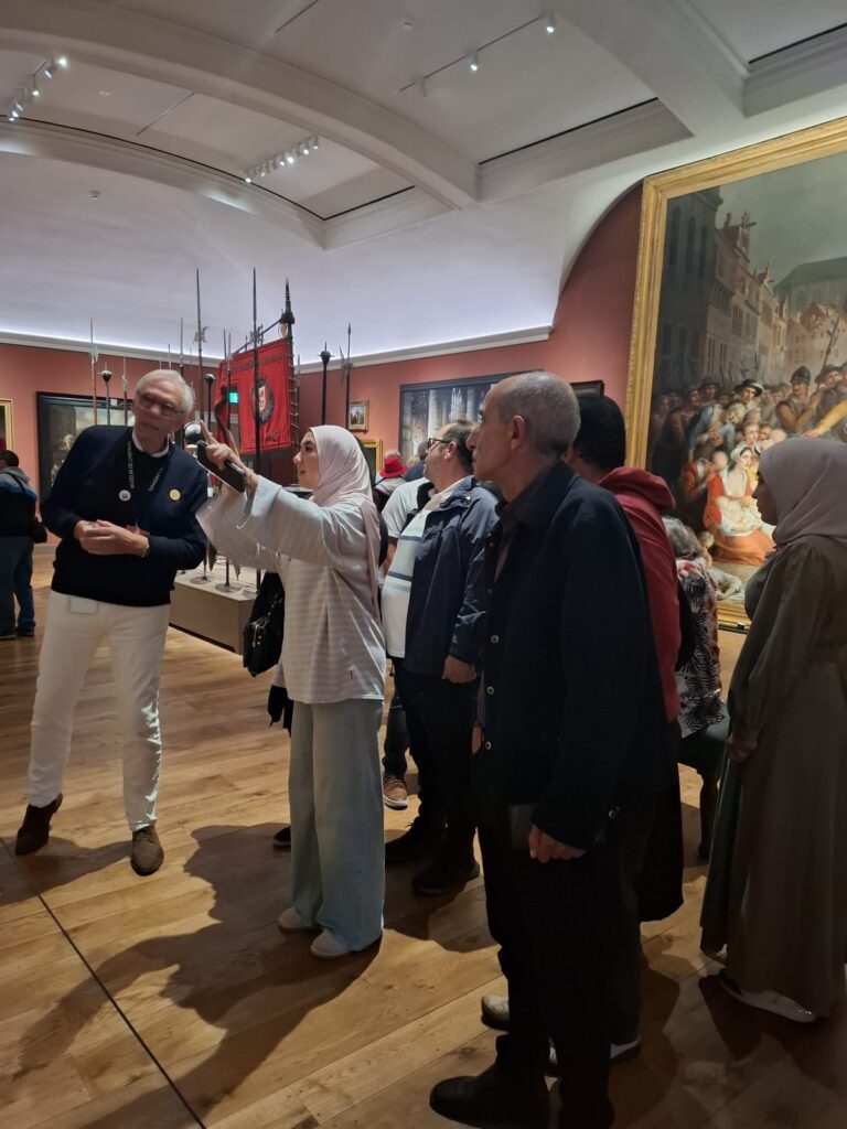 Een groep deelnemers van Stichting Sakiena krijgt uitleg in een zaal van Museum De Lakenhal. Een vrouw wijst naar boven, naast oude lansen, een rood vaandel en een groot schilderij in een gouden lijst.